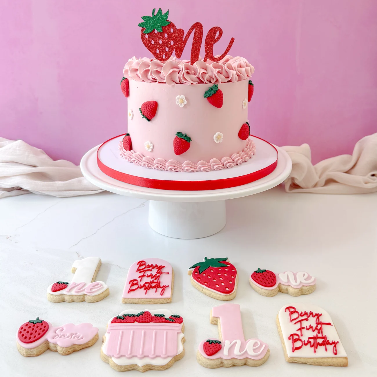 Pink strawberry first birthday cake with handmade strawberry decorations and matching cookies
